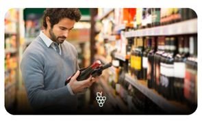 Homem analisando rótulo de vinho em prateleira de supermercado.